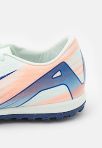 Sport shoe with white upper featuring a gradient of soft pink to light blue, textured fabric, and a dark blue sole with traction studs.