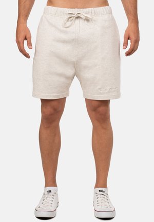 Mężczyzna w jasnobeżowych szortach na sznurek i białych niskich trampkach, stoi z nogami lekko rozstawionymi na jednolitym tle.