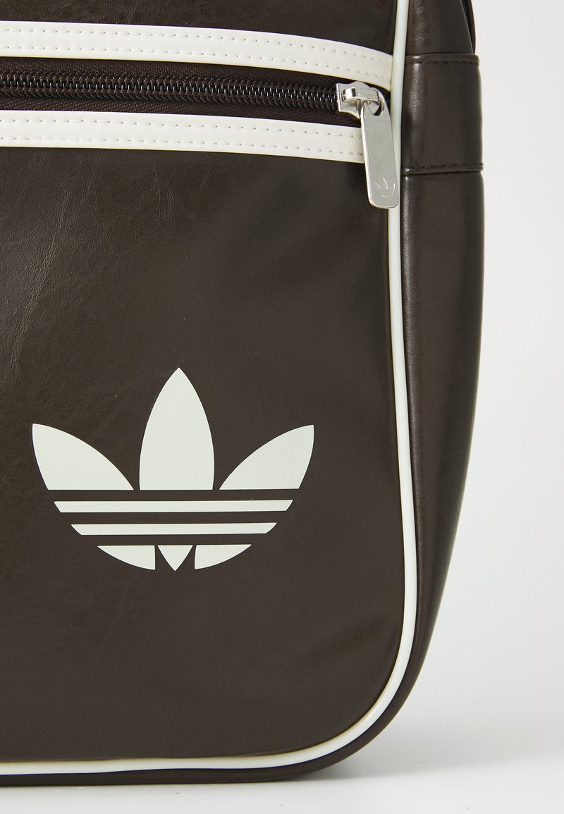 Sac en synthétique marron avec une fermeture éclair blanche, bordé de passepoil blanc, et un grand logo adidas blanc avec trois pétales et des rayures horizontales.