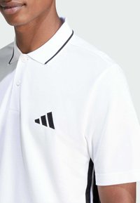 Witte poloshirt met een zwart geborduurd logo, zwarte accenten aan de zijkanten en contrasterende zwarte kraagafwerking, gemaakt van een gestructureerde stof.