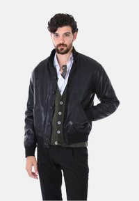 Giacca bomber in pelle nera con colletto e polsini a coste, dotata di chiusura frontale con bottoni e tasche laterali. Indossata sopra un cardigan verde.