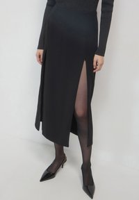 Jupe maxi noire avec fente avant, fabriquée en tissu lisse. Présente une taille haute et une fermeture éclair sur le côté. Portée avec des escarpins noirs à bout pointu et des collants transparents.