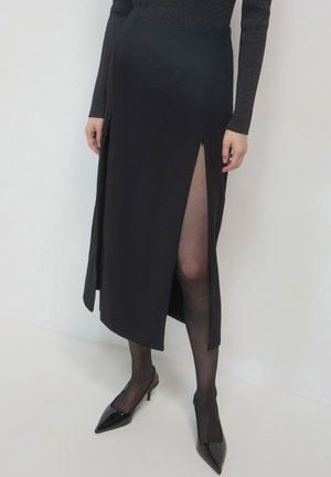 Gonna maxi nera con spacco frontale, realizzata in un tessuto morbido. Presenta una vita alta e una cerniera laterale. Indossata con tacchi a punta neri e collant trasparenti.