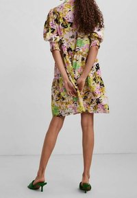 Bloemmotief jurk in pastelkleuren, met pofmouwen, een getailleerde taille en een gelaagde rok. Draagt groene open teen hakken.