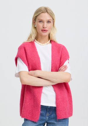 Kvinde med blond hår iført en pink ærmeløs cardigan over en hvid skjorte og blå jeans, stående med armene over kors.