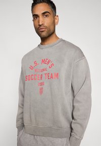 Pelēks džemperis ar apaļu kakla izgriezumu, ar sarkanu uzdruku "U.S. MEN'S NATIONAL SOCCER TEAM 2026" un logotipa elementiem. Mīksts, teksturēts audums.
