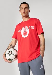 Rød bomulds t-shirt med "SCHWEIZ" trykt i hvid, båret med grå sportsbukser; holder en mønstret fodbold.