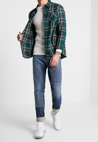 Vihreä-valkoraidallinen flanellipaita, kerroksittain ribbikudotun vaalean kermanvärisen neuleen päällä. Siniset farkut, joissa on kuviollisia kangaspäällysteitä ja käännetyt lahkeet. Valkoiset tennarit.