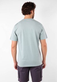 T-shirt bleu clair à manches courtes avec un col rond, texture lisse et un petit logo sur l'épaule gauche. Drapé de tissu visible.