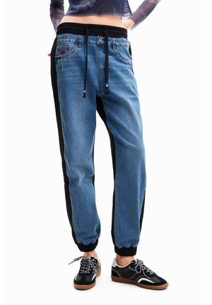 Persona con pantalones jogger de mezclilla azul con paneles laterales negros y zapatillas negras con suelas blancas y marrones, de pie sobre un fondo blanco.