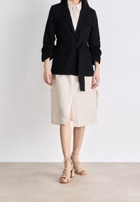 Žena oblečená v černém sako s opaskem přes béžové midi šaty na knoflíky a béžové sandály na blokovém podpatku se šněrováním, stojí na hladké podlaze.