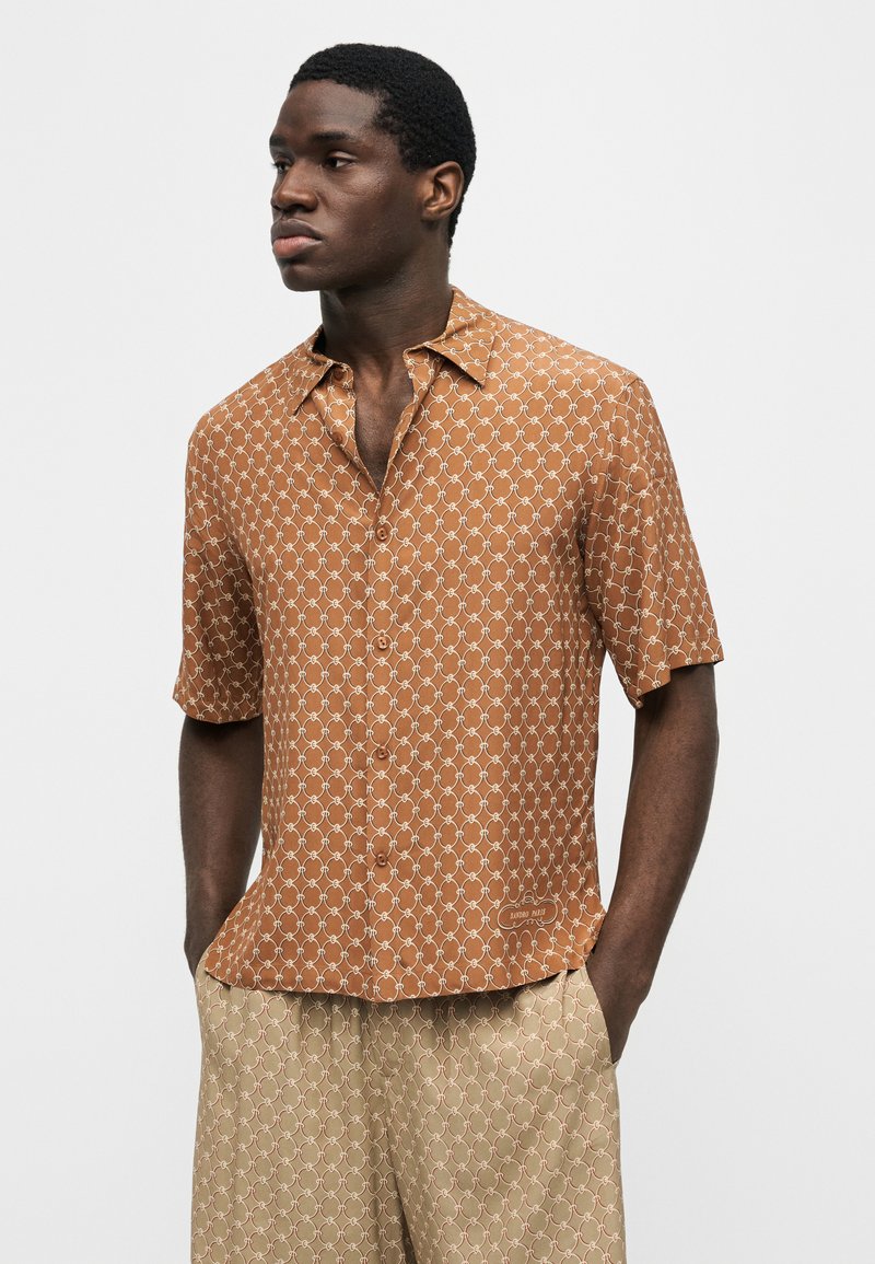 Mann trägt braunes kurzärmeliges Hemd mit weißem Kettengliedmuster und beigen Hosen mit ähnlichem Muster, Hände in den Taschen.