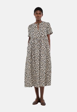 Femme portant une robe à manches courtes, longueur mollet, à motif léopard et des chaussures plates tressées marron, debout devant un fond uni.