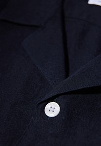 Ναυτικό πουκάμισο με κουμπιά και μαλακή υφή, που διαθέτει άσπρο κουμπί με τέσσερις τρύπες και κοφτό γιακά. Εστίαση στις λεπτομέρειες του υφάσματος και τη ραφή.