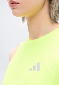 Gros plan d'une personne aux cheveux courts portant un haut sans manches jaune néon avec un logo gris à trois barres sur la poitrine.