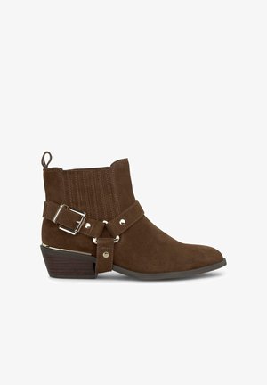 Bottine en suède marron avec un talon bas empilé, présente des coutures verticales, une sangle à boucle et des accents métalliques.