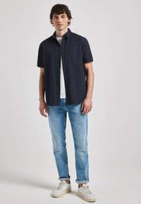 Camisa de botones de manga corta en color navy con un bolsillo en el pecho, combinada con vaqueros de corte claro y zapatillas blancas. Tejido suave y corte relajado.