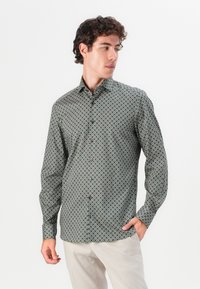 Dunkelgrüne Hemdjacke mit einem geometrischen Muster in Weiß und Hellgrün. Lange Ärmel und eine taillierte Passform. Braune Knöpfe.