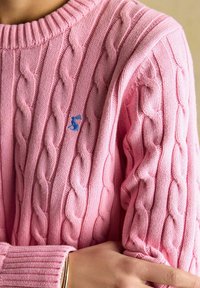 Zbliżenie różowego swetra w sploty ze wzorem, z niebieskim haftowanym logo królika, a na ramionach osoby założone skrzyżowane ręce z złotą bransoletką.