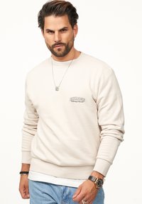 Beiger Sweatshirt mit langen Ärmeln, gerippten Bündchen und Rundhalsausschnitt. Verfügt über ein kleines Logo auf der Brust. Wird mit hellblauen Jeans getragen.