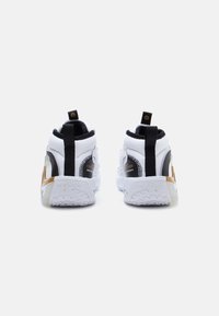 White athletic shoes featuring a black strap, textured accents, and a gold detail on the heel, with a thick, patterned rubber sole.
