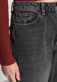 Černé džíny z denimu s klasickým designem pěti kapes, viditelnými švy a mírně vybledlou strukturou. Červený žebrovaný top s dlouhým rukávem je částečně viditelný.