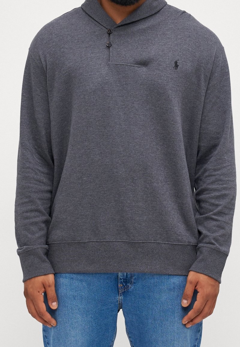 Homme portant un polo gris foncé à manches longues avec deux boutons et un petit logo noir brodé, associé à un jean bleu.