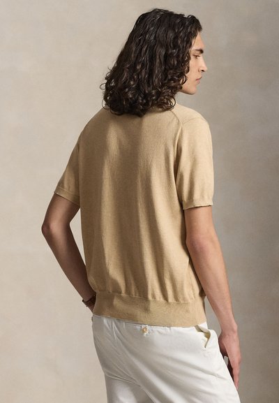 Άνδρας με σγουρά σκουρόχρωμα μαλλιά φορώντας μπεζ κοντομάνικο πουλόβερ και λευκό παντελόνι, φαίνεται από πίσω και πλαγίως.