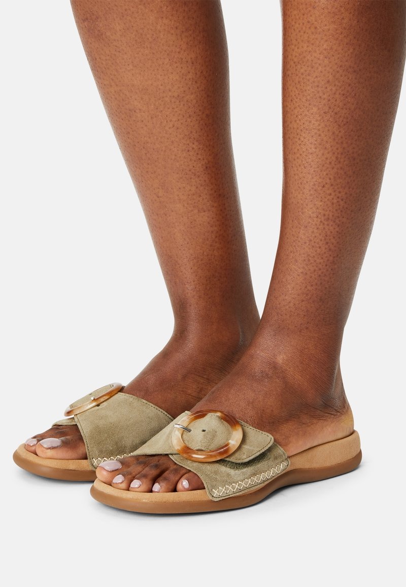 Pieds à la peau brune portant des sandales ouvertes en daim beige avec de grandes boucles rondes et des bords cousus sur un fond clair.