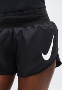 Μαύρα αθλητικά σορτς με ψηλή μέση και λευκό λογότυπο Nike στον δεξί μηρό, φορεμένα από άτομο με σκούρο δέρμα.