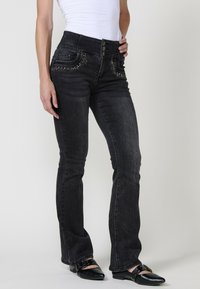 Črne flared jeans iz denima s dekorativnimi žebljički na sprednjih žepih, visokim pasom z zapiranjem na tri gumbe in gladko, teksturirano površino.