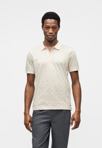 Μπεζ πόλο μπλούζα με διακριτικό υφασμάτινο μοτίβο, απαλό ύφασμα και σχεδιασμό με γιακά. Σε συνδυασμό με γκρι παντελόνια, εφαρμοστά μανίκια και τρία κουμπιά.