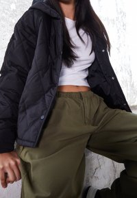 Schwarze gesteppte Jacke mit Kapuze, weißes kurzes Hemd und lockere olivgrüne Hose. Glatte Texturen und kontrastreiche Farben. Lässige Passform.