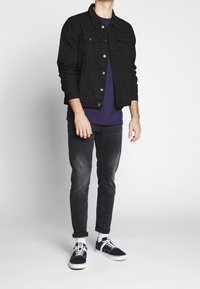 Mies, joka pitää yllä mustaa farkkutakkia, tumman violettia paitaa, haalistuneita mustia farkkuja, joita on käännetty nilkoista, valkoisia sukkia ja mustia tennareita, seisoo yksinkertaisella taustalla.