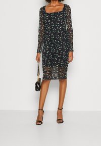 Zwarte mesh jurk met lange mouwen, bloemmotieven in groen en wit, geplooide details, vierkante halslijn en lengte tot halverwege de kuit.