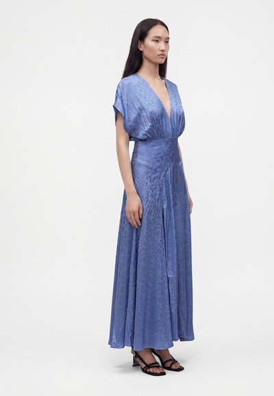 Femme portant une longue robe bleue fluide avec un profond décolleté en V et des manches courtes, associée à des talons ouverts noirs, debout sur un fond blanc.