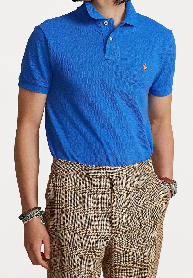 Homme portant un polo bleu à manches courtes rentré dans un pantalon marron à carreaux, avec des bracelets au poignet gauche et une montre au poignet droit.