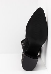 Semelle d'une botte noire à bout pointu, inclinée contre un mur blanc, montrant le talon texturé ainsi qu'une partie de la tige en daim clouté avec une fermeture éclair latérale.