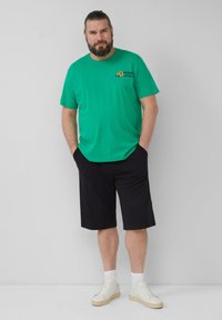 Groene katoenen t-shirt met logo, zwarte shorts, witte sneakers en sokken. Model staat met de handen in de zakken tegen een effen achtergrond.
