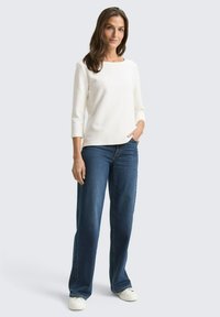 Weißes, geripptes Oberteil mit drei Viertelärmel, kombiniert mit weit geschnittenen dunkelblauen Jeans und weißen Sneakers. Glatte Textur und lockere Passform.