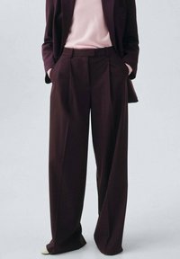 Wide-leg trousers in deep burgundy fabric, featuring a high waist, pleats, and side pockets. Worn with a light pink top.