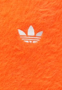 Ryški oranžinis, pūkuotas tekstilės gaminys su ryškiu baltu siuvinėtu Adidas trefoil logotipu ir trimis juostelėmis po juo.