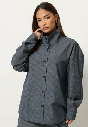 Femme aux longs cheveux foncés portant une chemise grise à carreaux oversize avec un col montant, regardant la caméra, main près du visage.