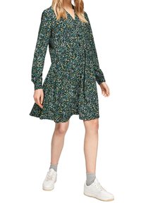 Blommig klänning med långa ärmar, krage, mörkblå bas, mönster i grönt, gult och vitt; utsvängt kjol; kombinerad med vita sneakers.