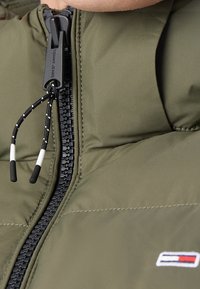 Vadderad olivgrön jacka med dragkedja, med en svart dragkedja med reflexdetaljer och en liten broderad logotyp på bröstet.