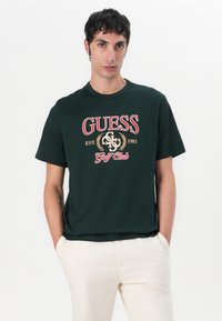 Camiseta de algodón verde oscuro con el logo "GUESS Golf Club" en rojo y dorado, con un corte relajado y escote redondo. Los detalles incluyen el año "1981."