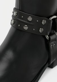 Μαύρο δερμάτινο μποτάκι αστραγάλου με φαρδιά λωρίδα διακοσμημένη με μεταλλικά τρουκς και λεπτομέρειες, με ομαλή υφή και στρογγυλεμένη μύτη.