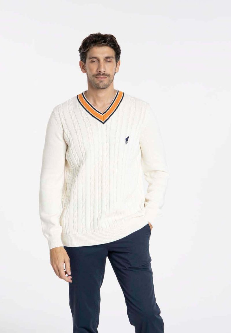 Pull en tricot torsadé crème avec un col en V, doté d'un col rayé orange et marine, porté avec un pantalon marine. Logo discret sur la poitrine.