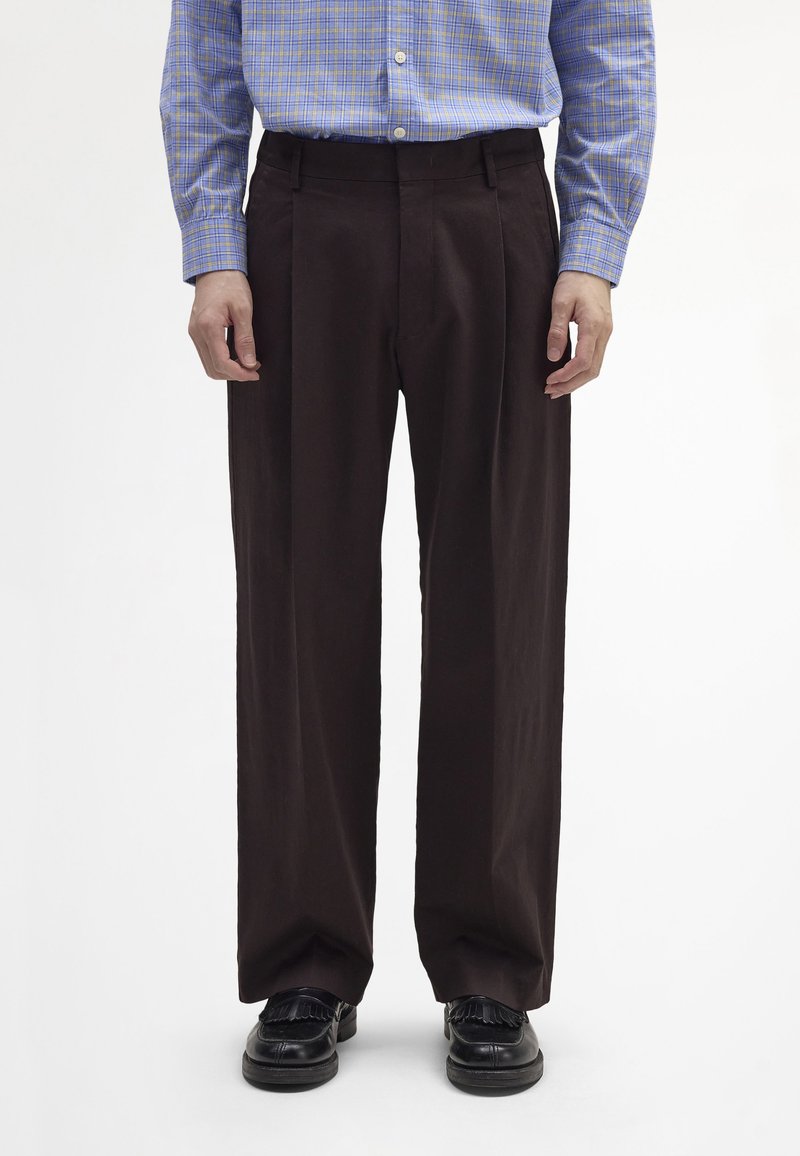 Barna, bő szárú nadrág elülső redőkkel, sima anyagból készült. Kék kockás inggel és fekete cipővel viselve.