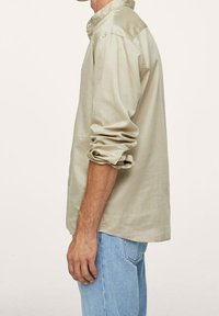 Jasno beżowa koszula zapinana na guziki, z zagiętym kołnierzykiem, podwiniętymi rękawami i miękką fakturą, w zestawieniu z jasnoniebieskimi dżinsami.